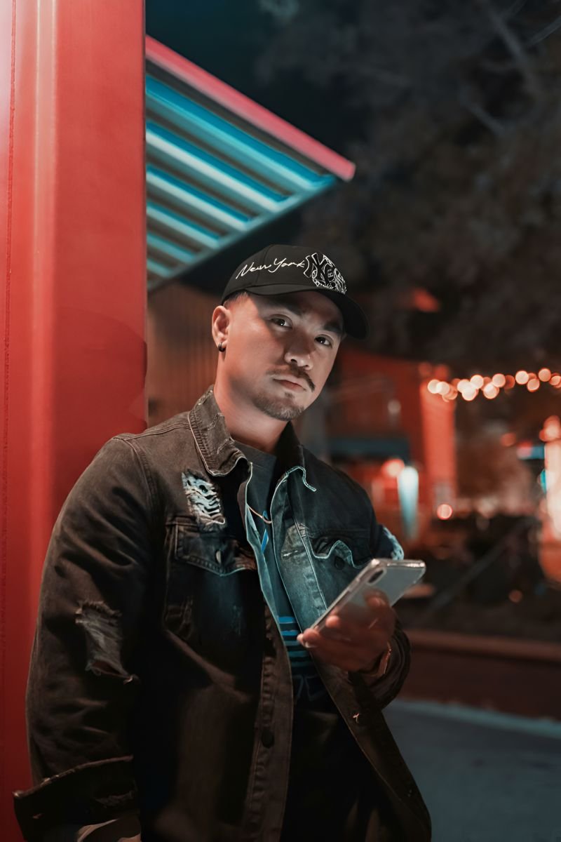 Hombre con chaqueta de mezclilla rasgada y gorra negra DANDY HATS x NY Favoritas “NY SKELETON”, posando de noche en un entorno urbano iluminado, mientras sostiene un celular.