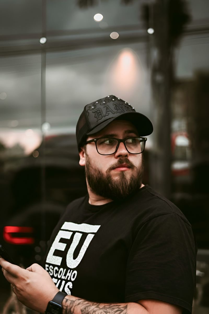 Hombre con barba y gafas usando una gorra negra Dandy Did This — What Would Dandy Do?, con detalles metálicos y bordado en alto relieve, mientras sostiene un celular frente a una fachada urbana.