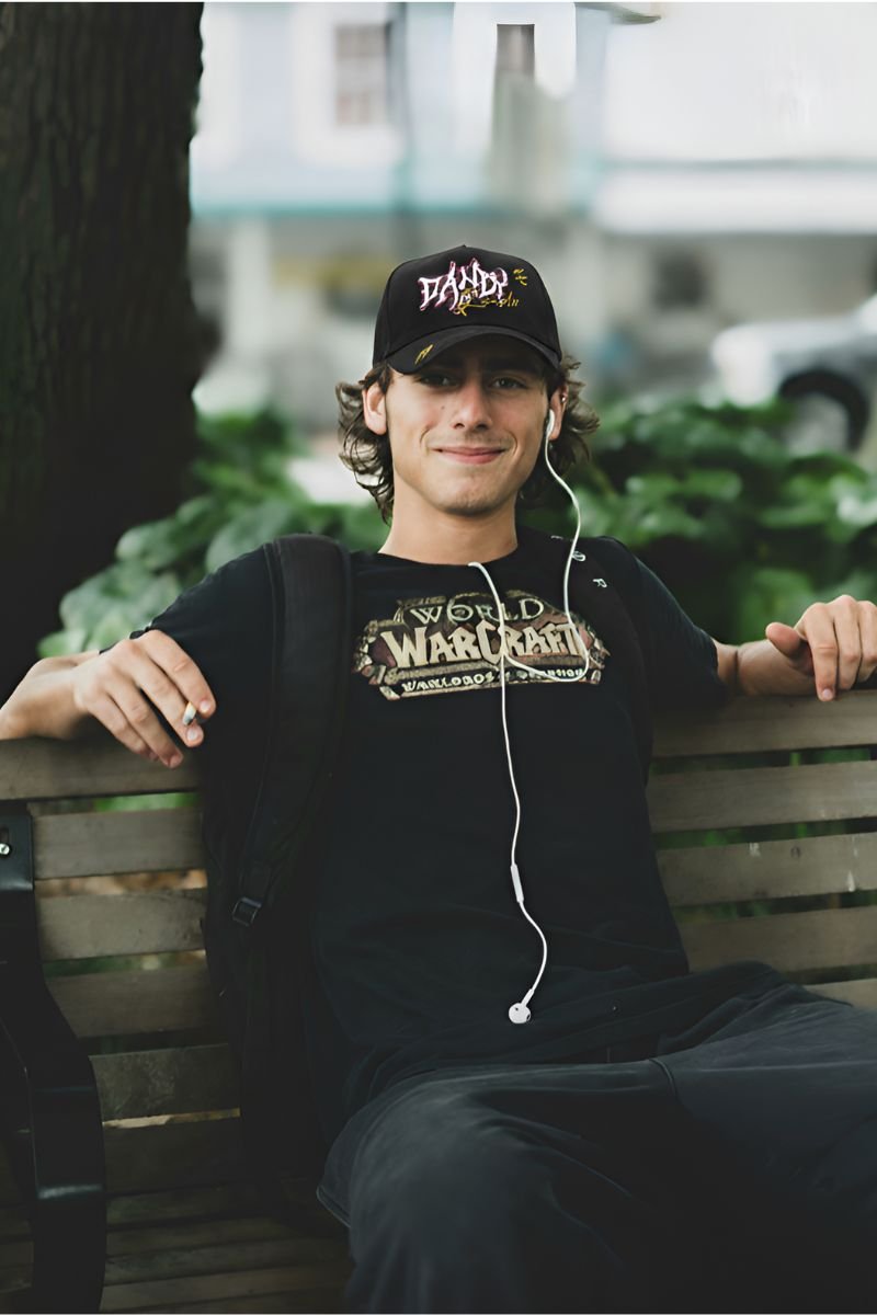 Joven sentado en una banca de parque con auriculares puestos, usando una gorra negra Dandy Hats “Records Black” y camiseta de World of Warcraft, con una expresión relajada y amistosa.