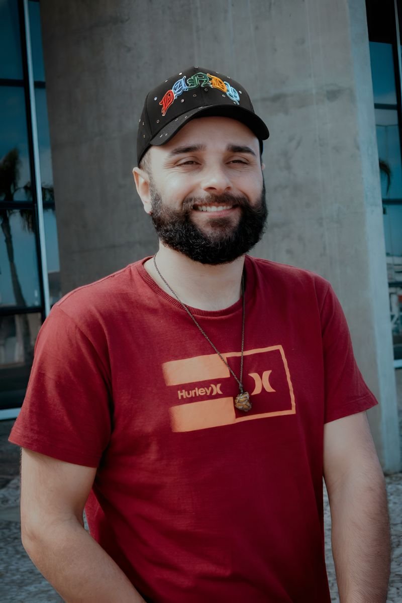 Hombre sonriente con barba usando una gorra negra DANDY HATS X 10º Aniversario con detalles multicolor, camiseta roja Hurley y collar con colgante, posando frente a un edificio moderno.