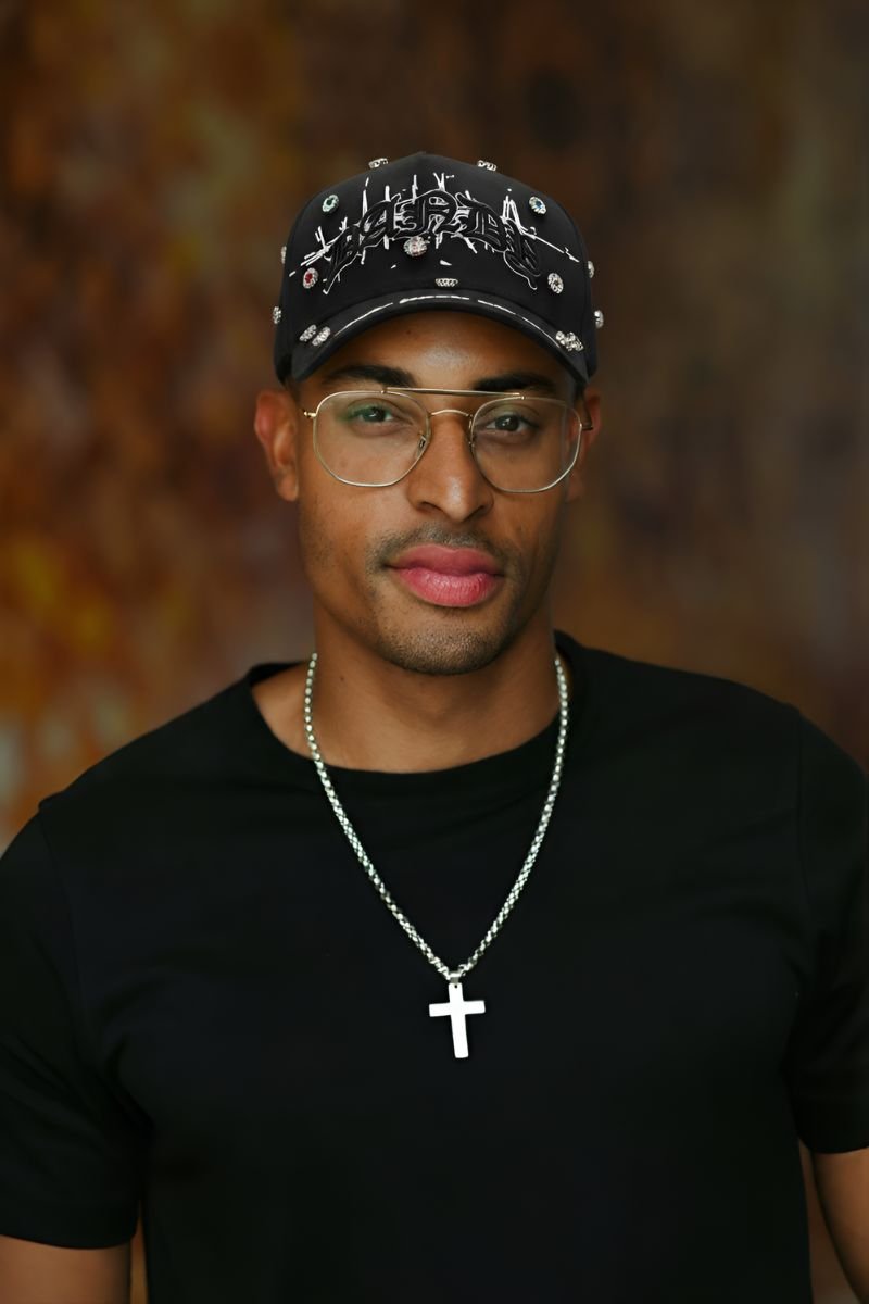 Hombre posando con una gorra negra Dandy Hats x Lil Baby “Bling” decorada con pedrería brillante, gafas doradas y camiseta negra, usando un collar con cruz plateada sobre fondo difuminado cálido.