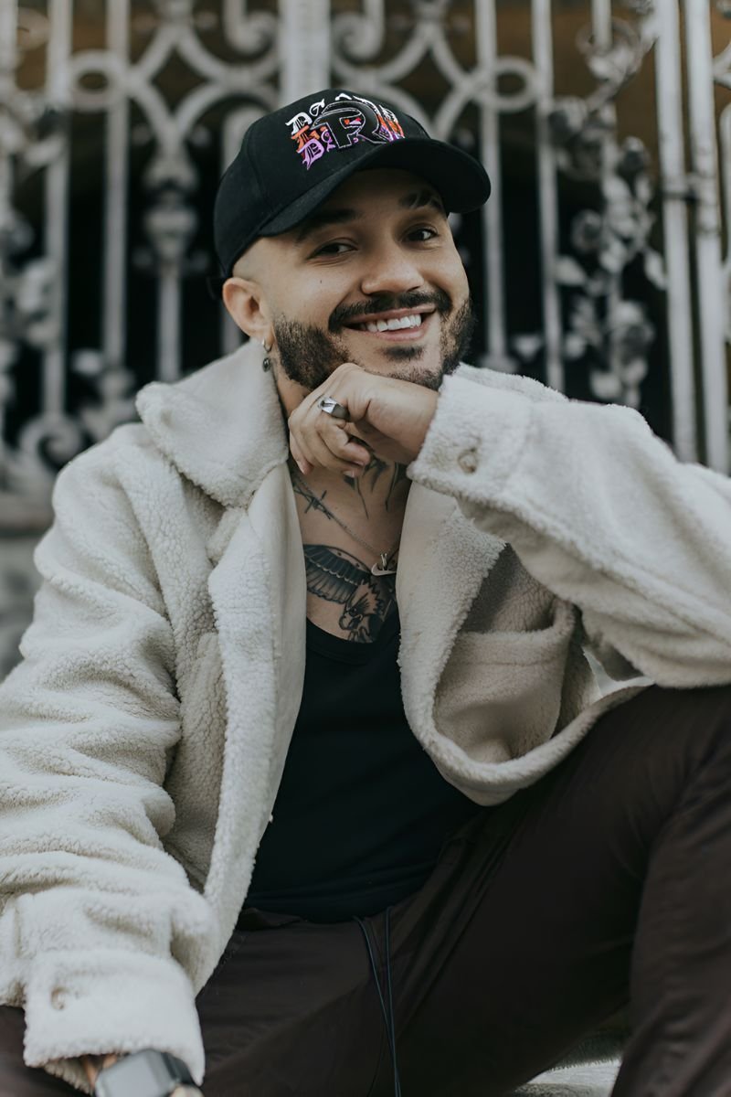 Hombre sonriente con gorra negra “FR DANDY” de la colaboración DANDY HATS x Fuerza Regida, usando chaqueta de borrego clara y camiseta negra, posando frente a una reja decorativa.