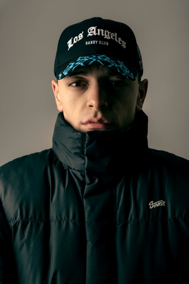Hombre joven usando gorra negra “LA DANDY CLUB” de la colaboración Dandy Hats x LA Favoritas, con chaqueta acolchada negra y fondo neutro.