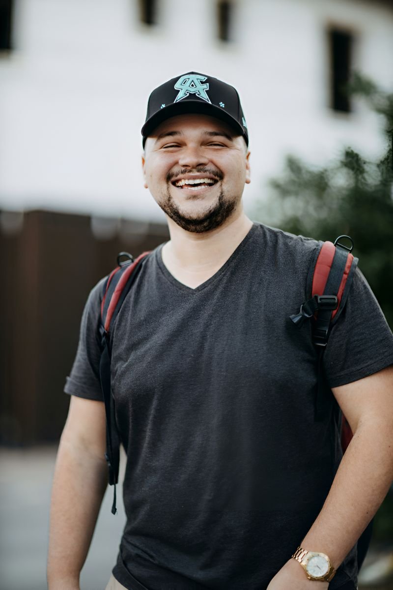Hombre joven sonriente con barba ligera, usando una gorra negra con diseño verde claro y una camiseta gris oscuro, cargando una mochila con correas rojas.