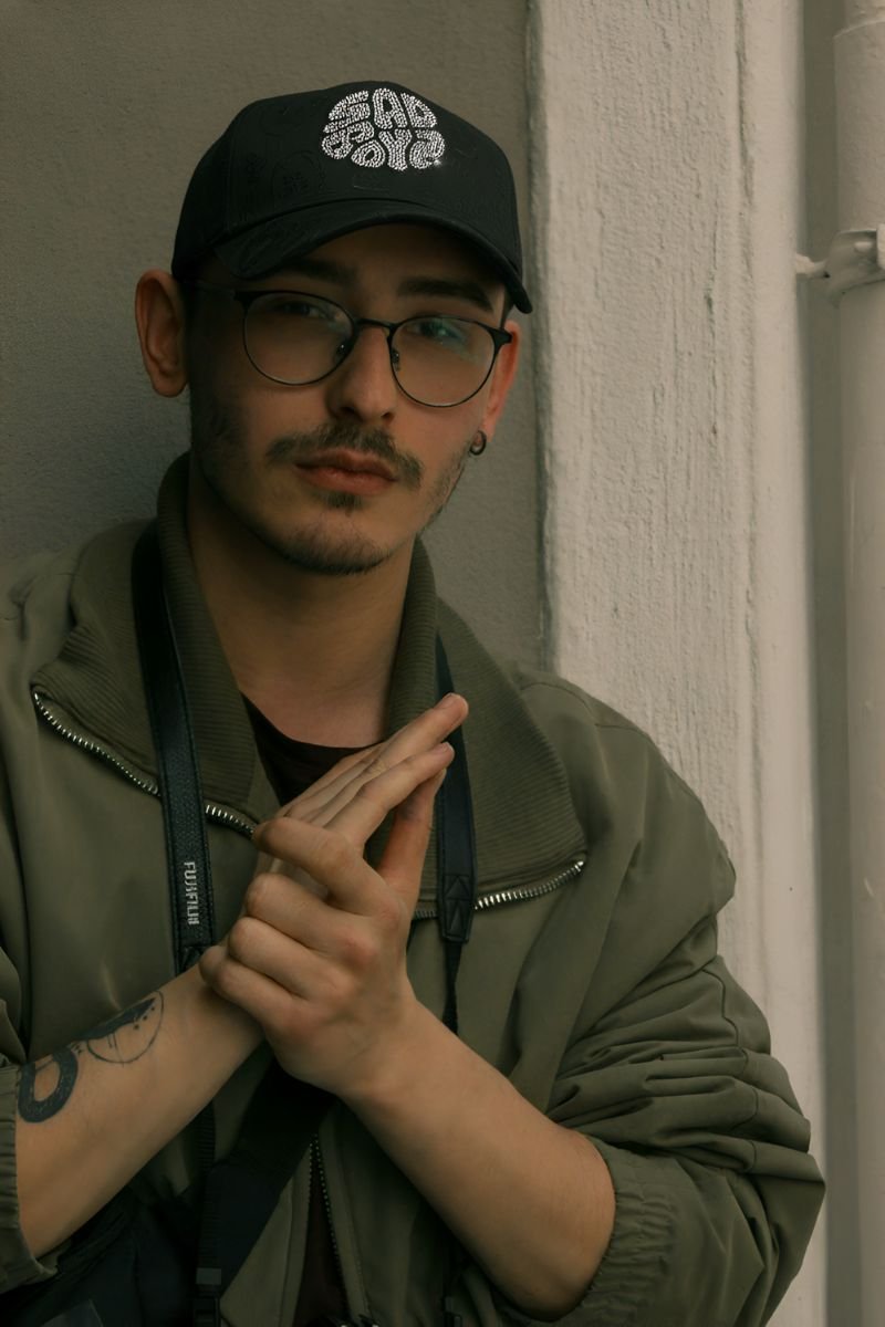 Hombre joven con gafas redondas, gorra negra con diseño brillante, chaqueta verde oliva y correa de cámara colgando del cuello, posando frente a una pared clara.