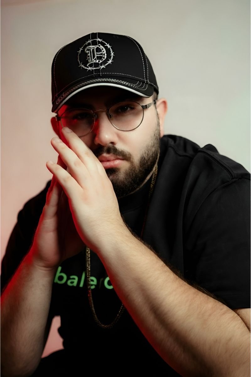 Hombre con gafas y camiseta negra posando con las manos juntas, usando una gorra negra Rodeo — DandyWood: Edición Exclusiva Sneaker Fever MX con diseño de alambre de púas y una “D” bordada.